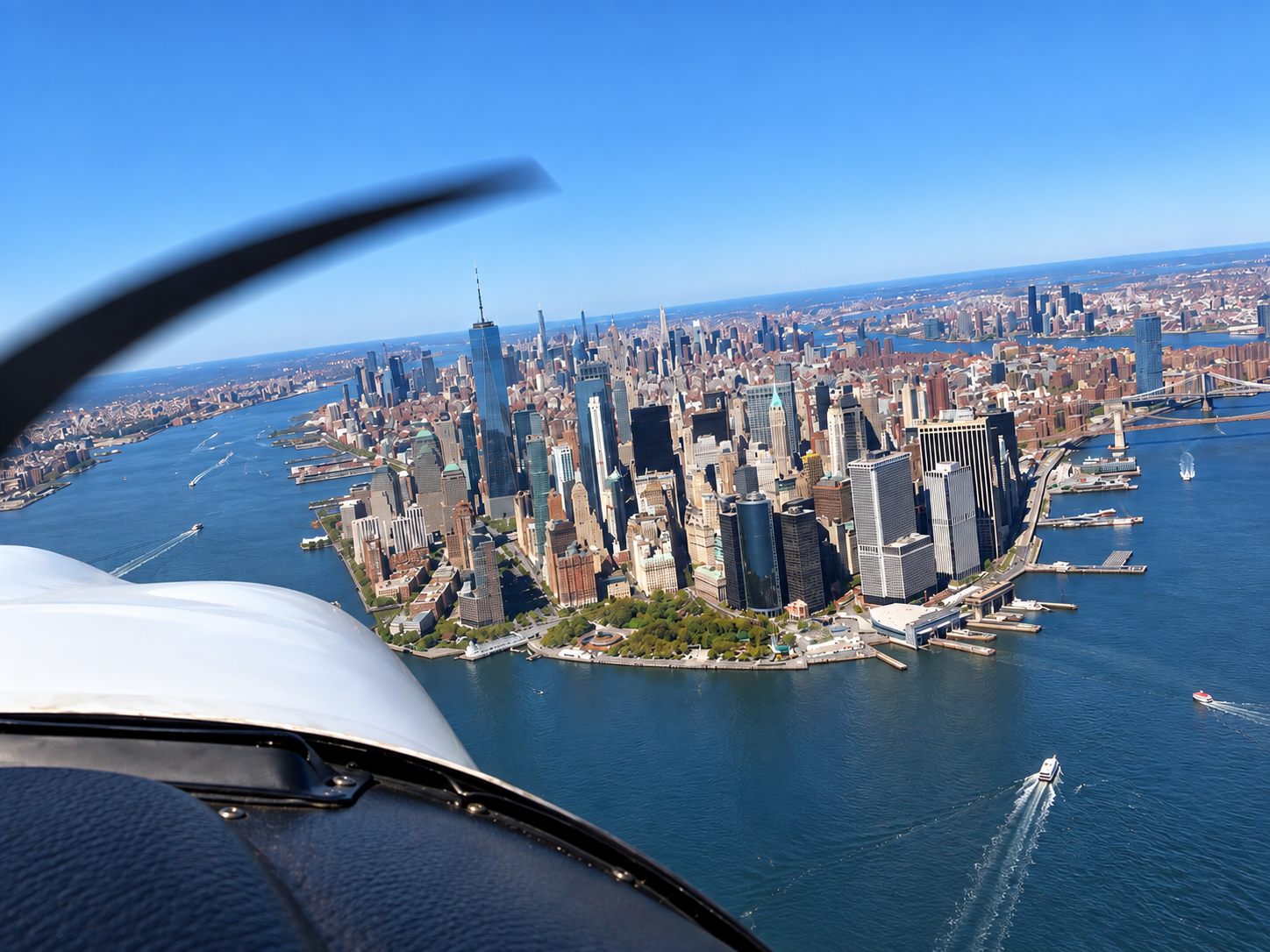 Aerial view of Manhattan, New York City, taken from a small plane, showing the skyline with tall skyscrapers, the harbor with boats and ships, and Brooklyn and New Jersey in the distance.