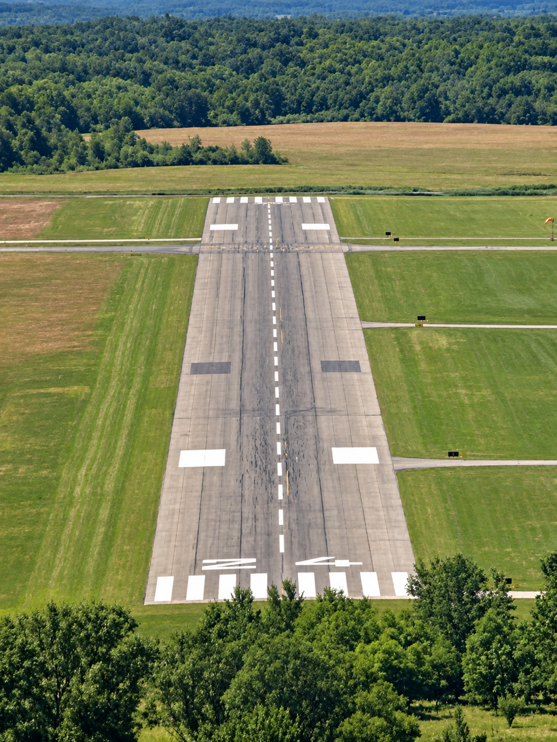 Aerial view of a small, empty runway with grassy fields and forested hills in the background.