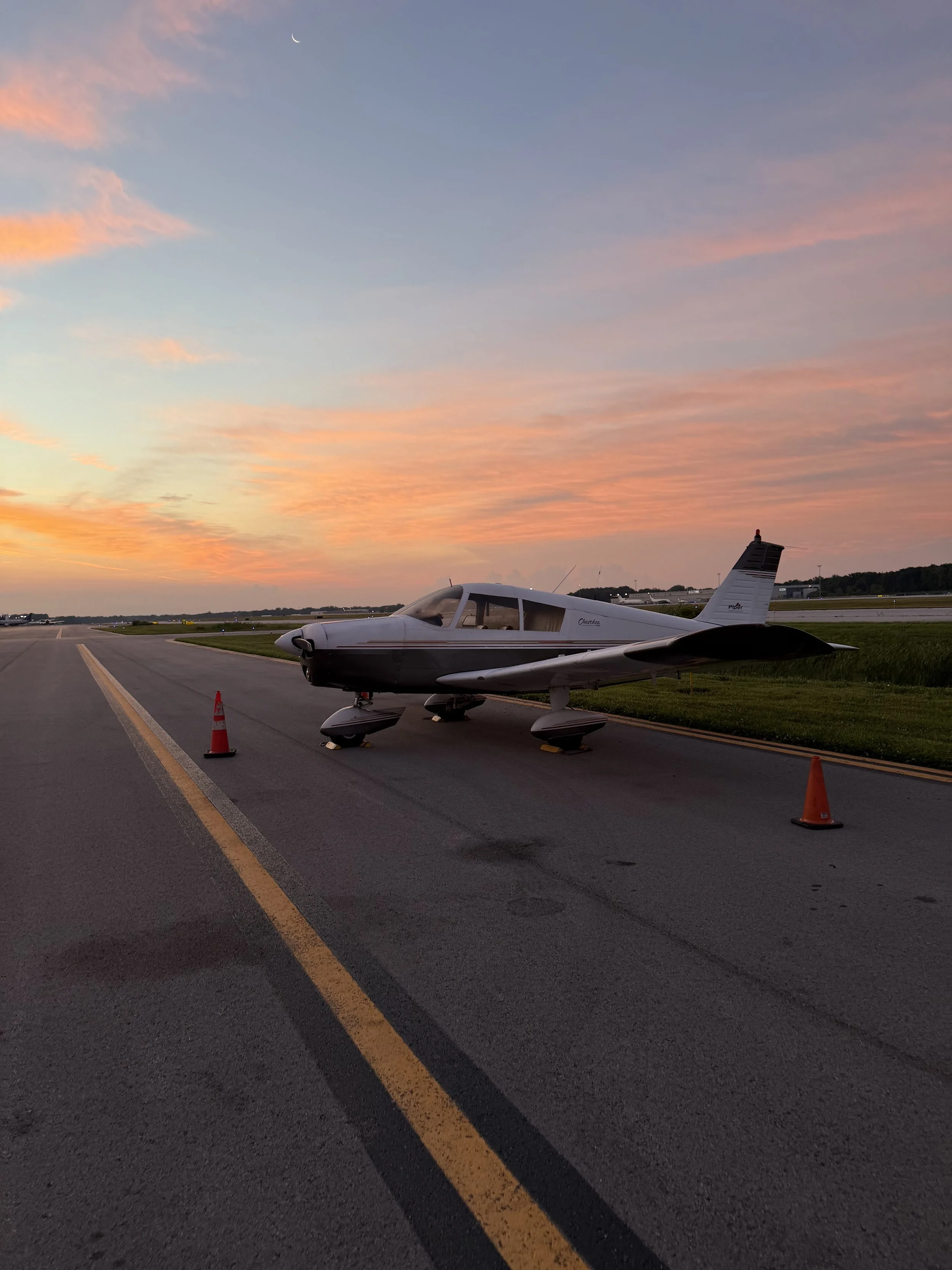 Small private jet on runway during sunset, with orange and pink clouds and a crescent moon in the sky.