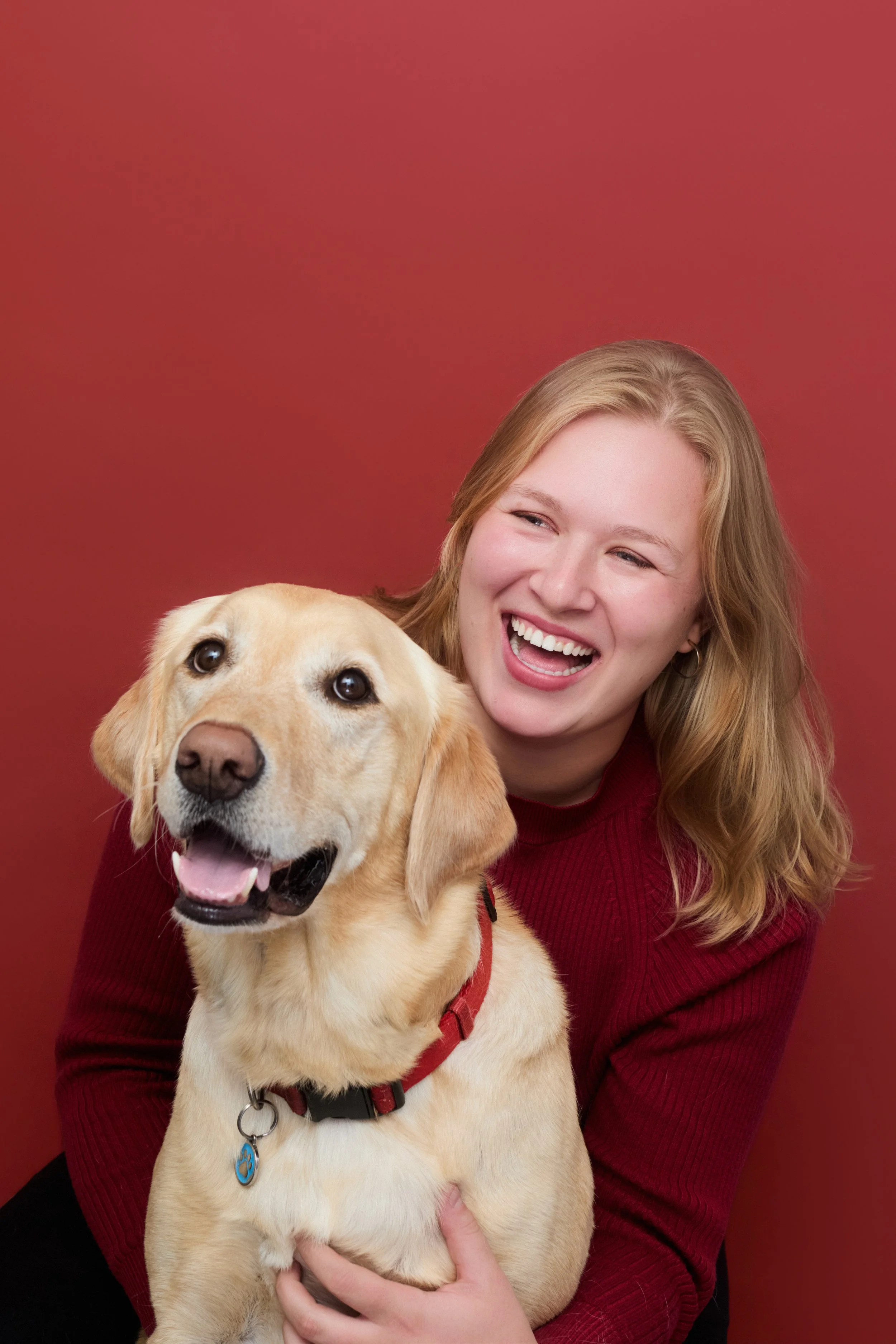 A woman with long wavy red hair, smiling brightly, holding a happy golden retriever dog with a red collar, set against a plain red background.