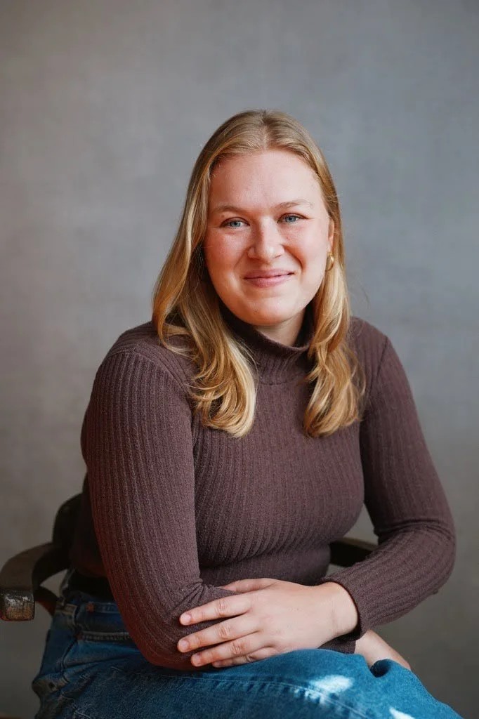 A young woman with blonde hair and blue eyes, wearing a maroon ribbed turtleneck sweater, sitting on a wooden chair, smiling, with a neutral grey background.