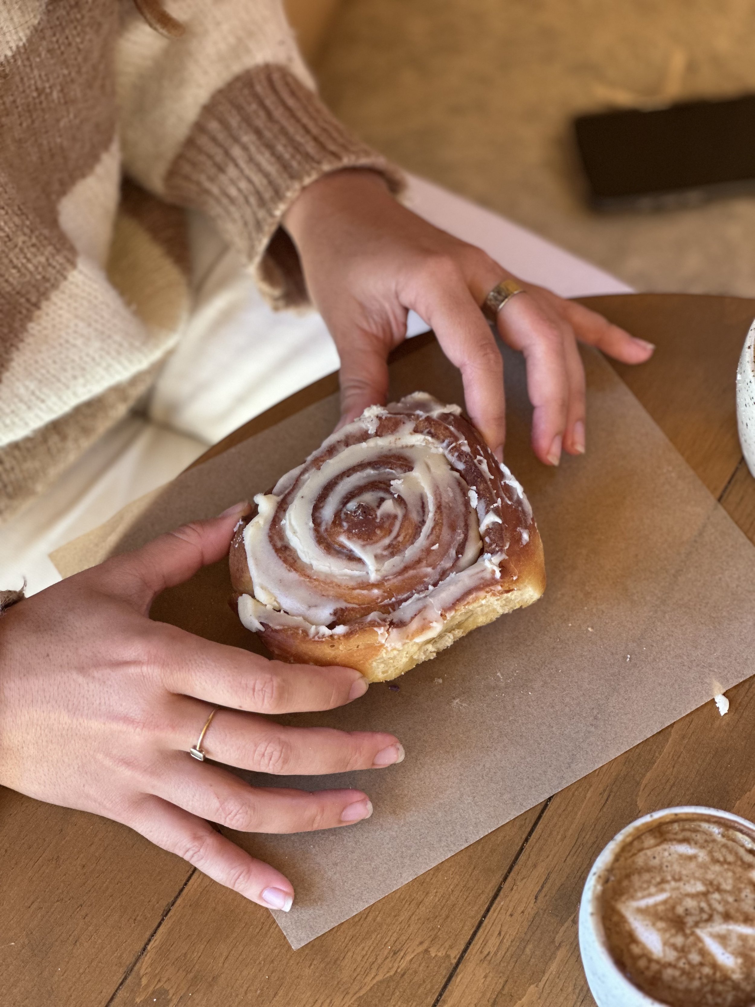 Person holding a cinnamon roll with white icing, sitting at a wooden table with a cup of coffee nearby. The person is wearing a beige and brown striped sweater.