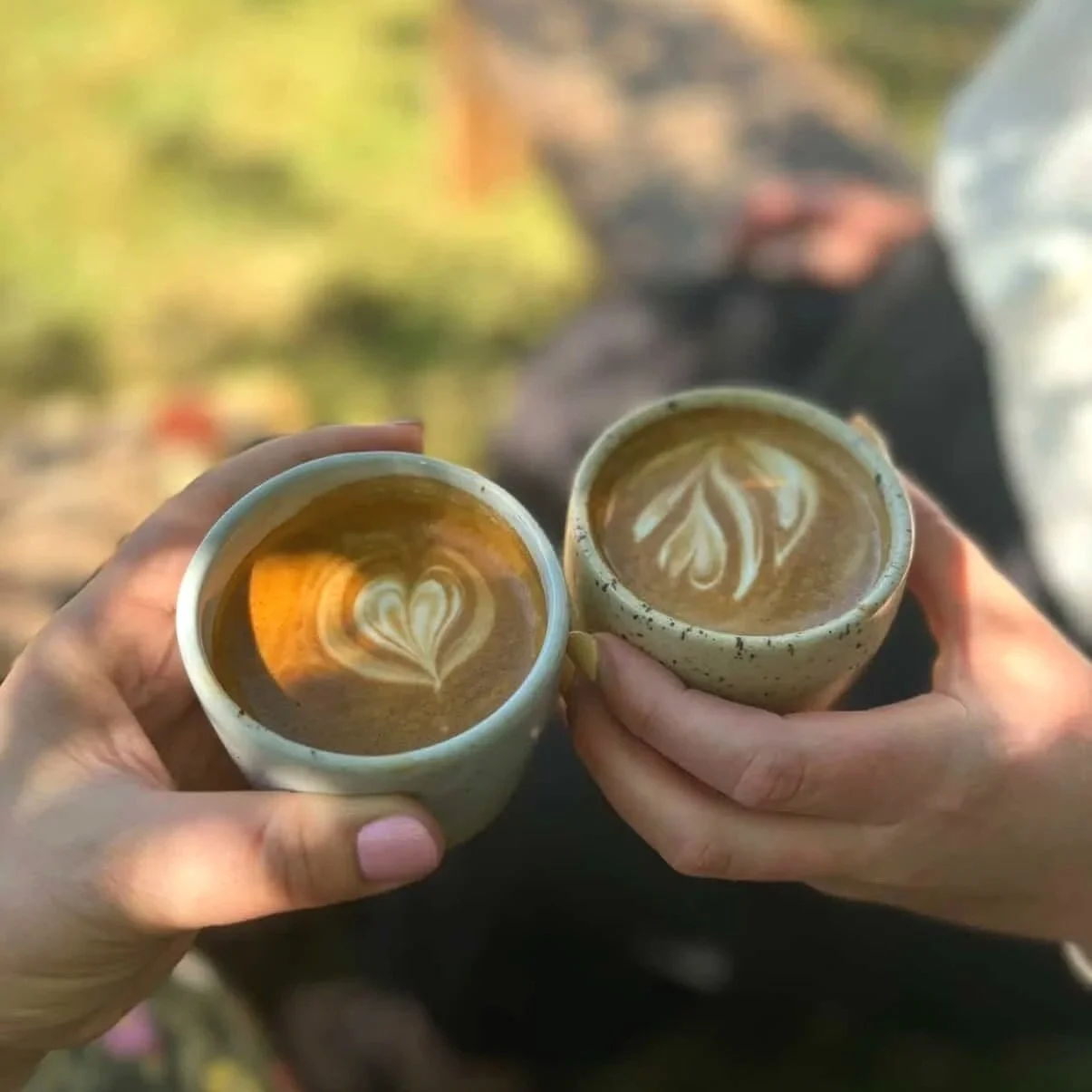 Two cups of coffee with latte art hearts, held by two people during daytime.