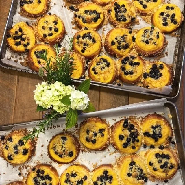 Wooden table with two baking sheets of baked peach desserts topped with blackberries and crumbles, garnished with white and green flowers.