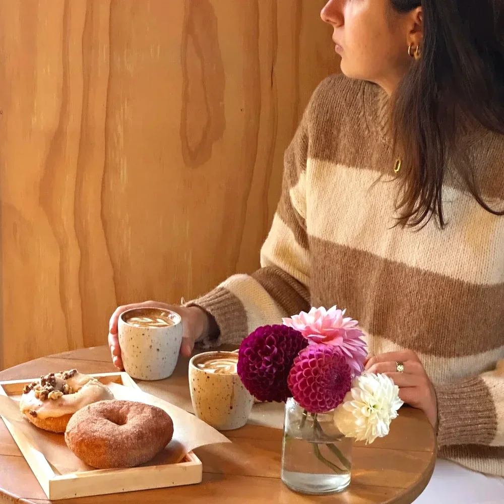 A woman wearing a brown and beige striped sweater sitting at a wooden table with two cups of coffee, a small vase with pink, dark red, and white flowers, and a tray of donuts with toppings.