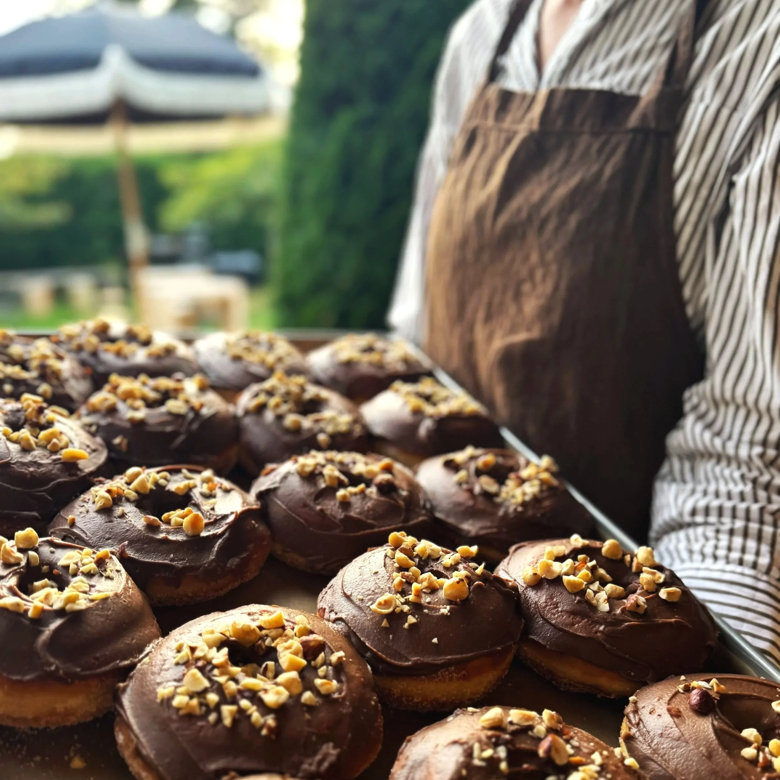 Close-up of assorted chocolate brioche topped with streusel, arranged on a tray with a person wearing a STABLE apron in a garden.