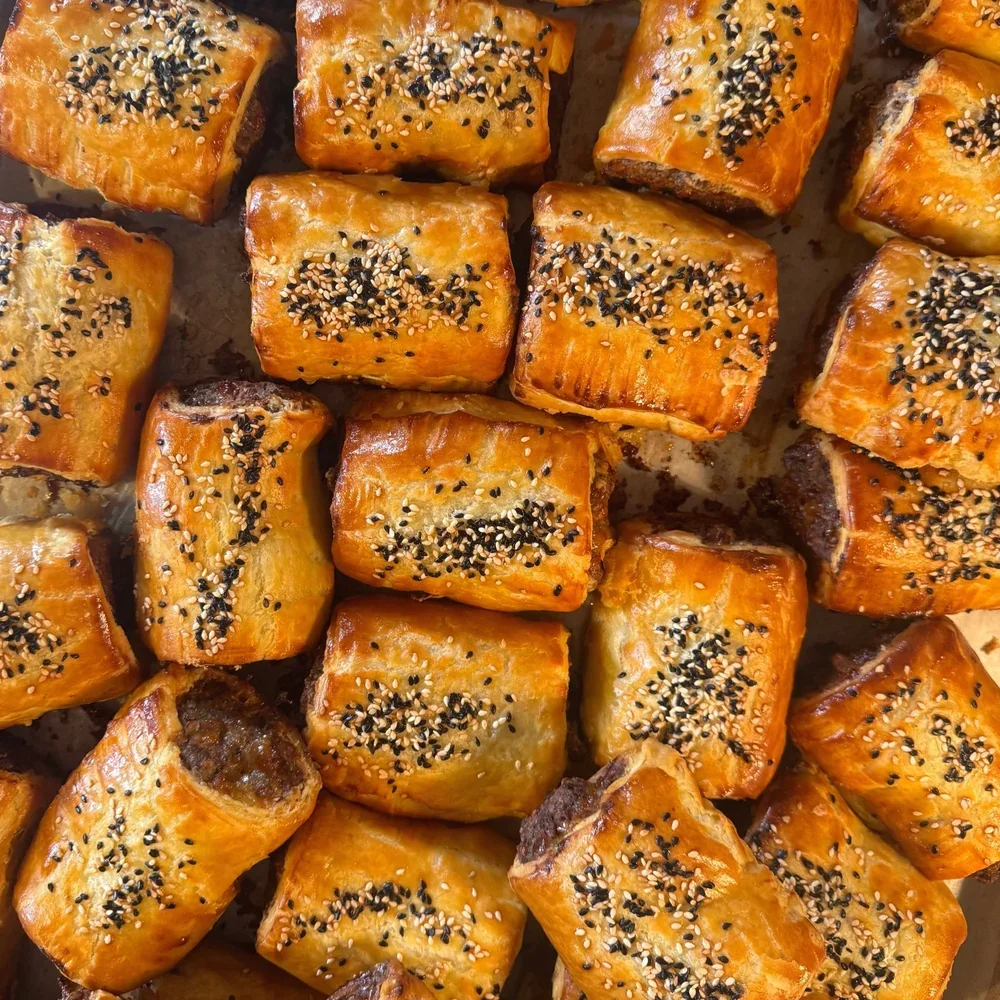 Golden-brown sausage rolls topped with sesame seeds on a baking sheet.