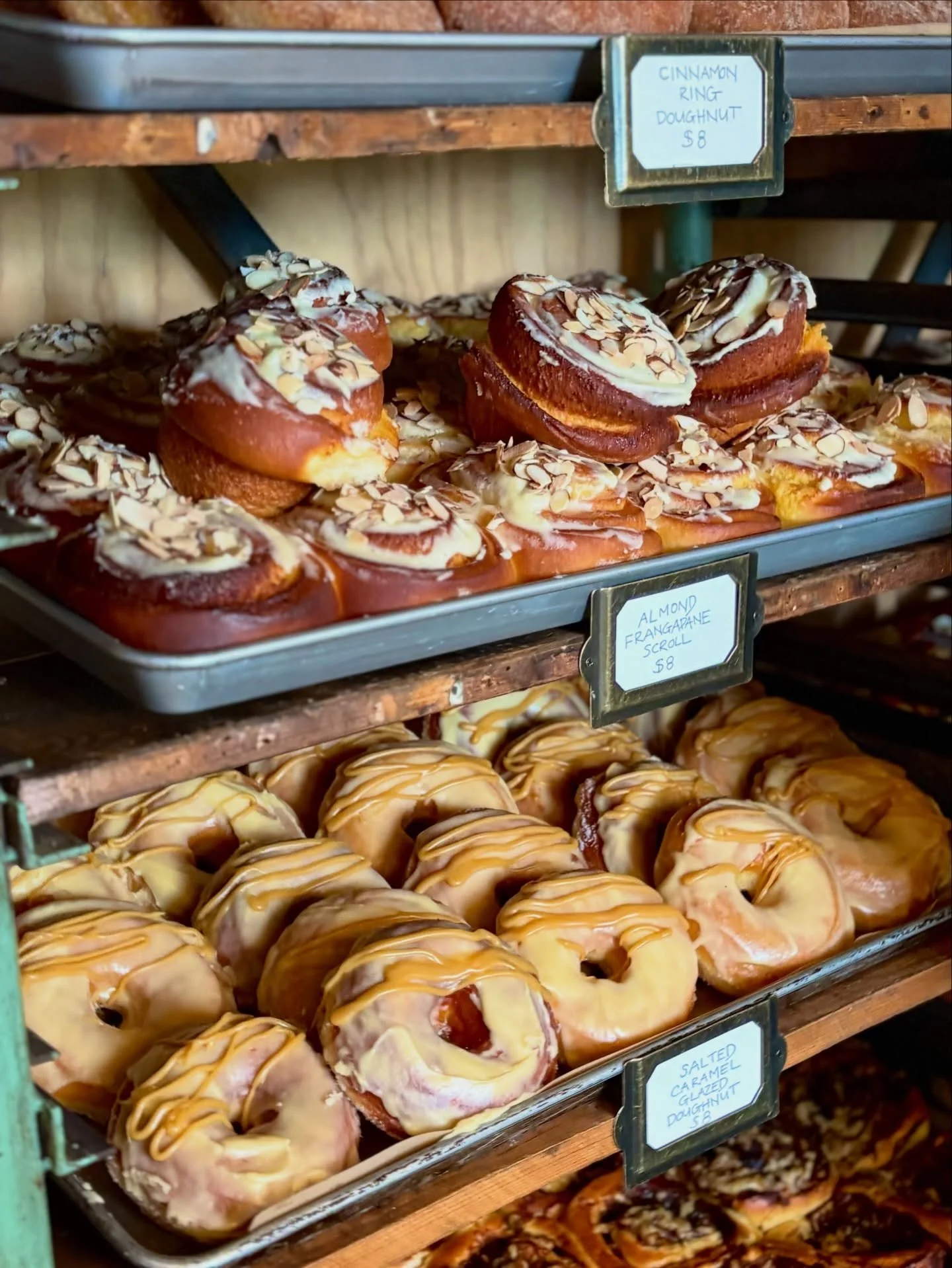 Display shelf with freshly baked donuts topped with almond slices, caramel glaze, and cinnamon rolls labeled cinnamon ring doughnut and salted caramel glazed doughnut, priced at $8.