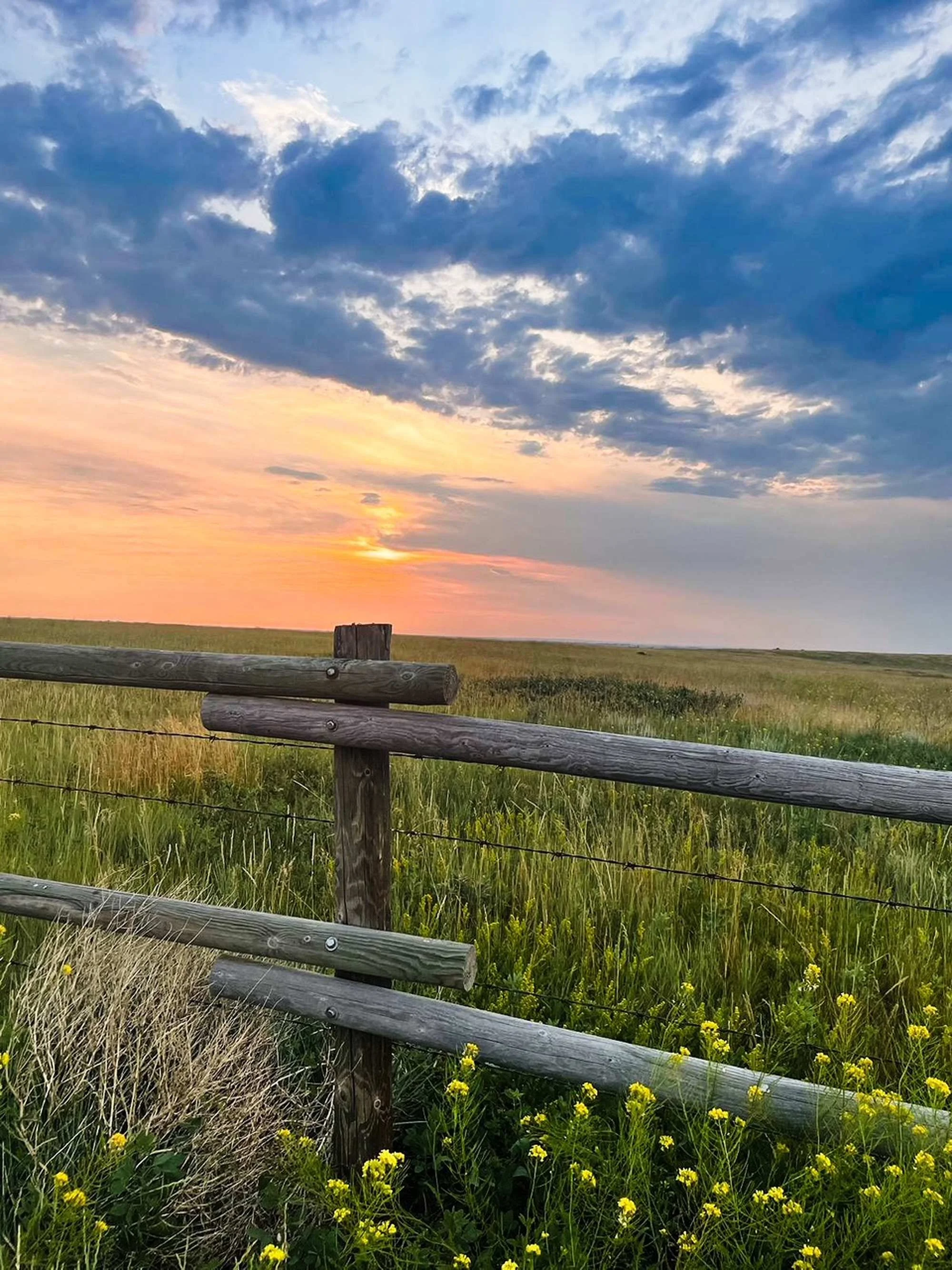 A scenic view of a sunset over a grassy field with a wooden fence in the foreground and a partly cloudy sky.