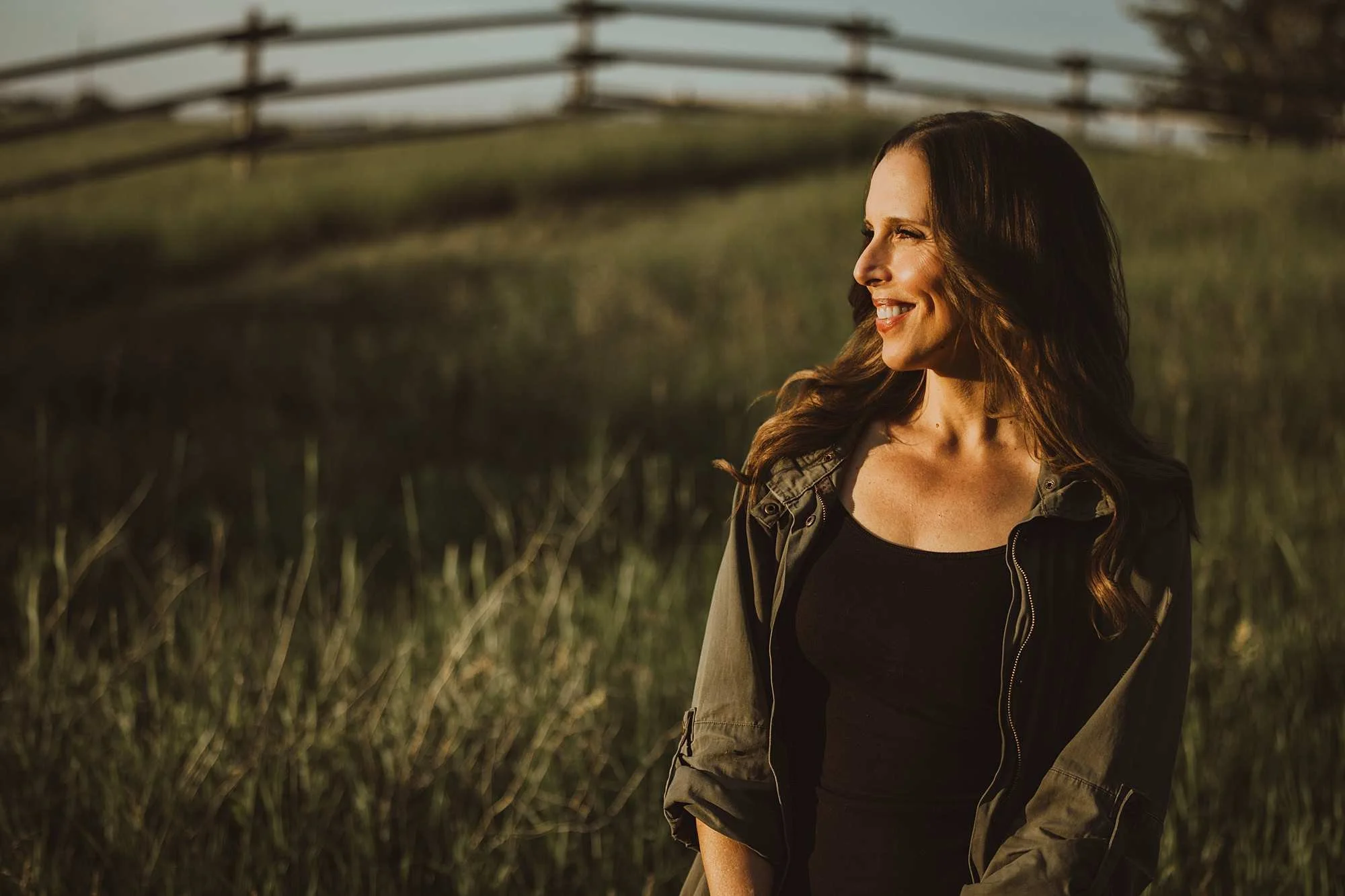 A woman with long brown hair and a bright smile standing outdoors in a field of tall grass, wearing a green jacket and black top, during golden hour in the late afternoon or early evening.