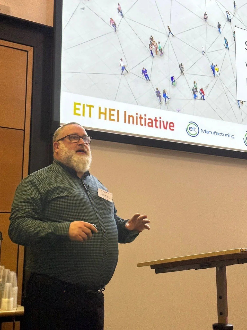 A man with glasses and a beard giving a presentation in front of a screen displaying a slide titled 'EIT HEI Initiative'. The slide shows a network of people and logos related to EIT Manufacturing.