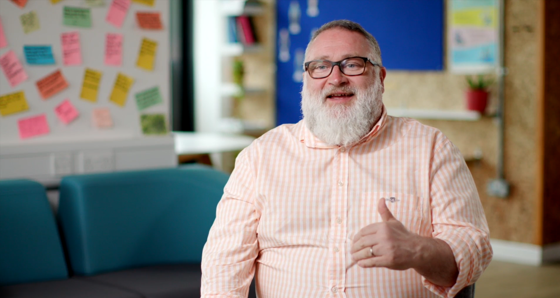 A happy man with a beard and glasses wearing a peach plaid shirt, sitting in a colorful office or classroom environment with a bulletin board and pink, yellow, and green notes in the background.