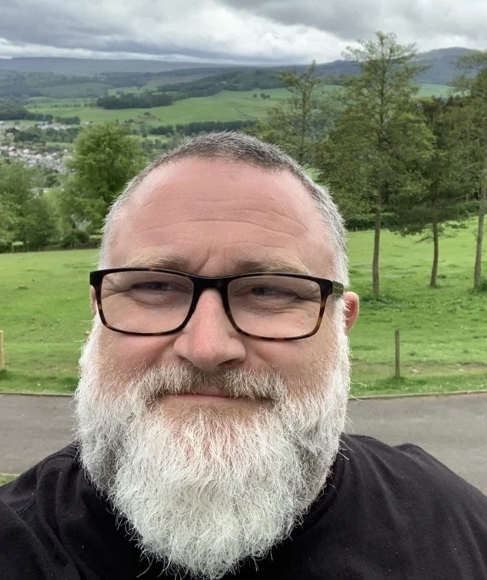 A man with glasses and a white beard taking a selfie outdoors with a grassy field, trees, and rolling hills in the background under cloudy skies.