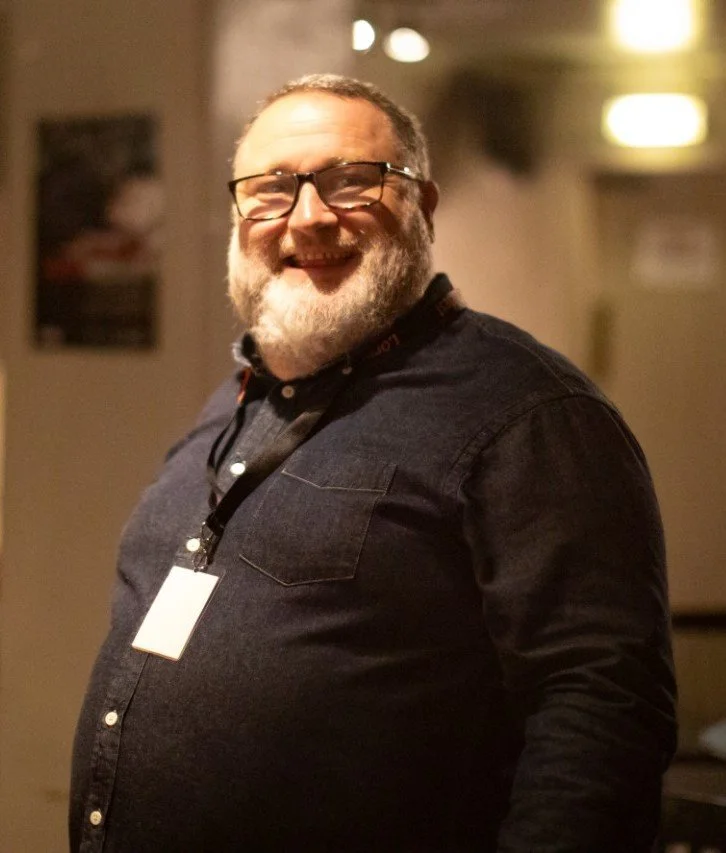 A smiling man with glasses, a beard, and a nametag, wearing a dark shirt, standing indoors in a warmly lit room.