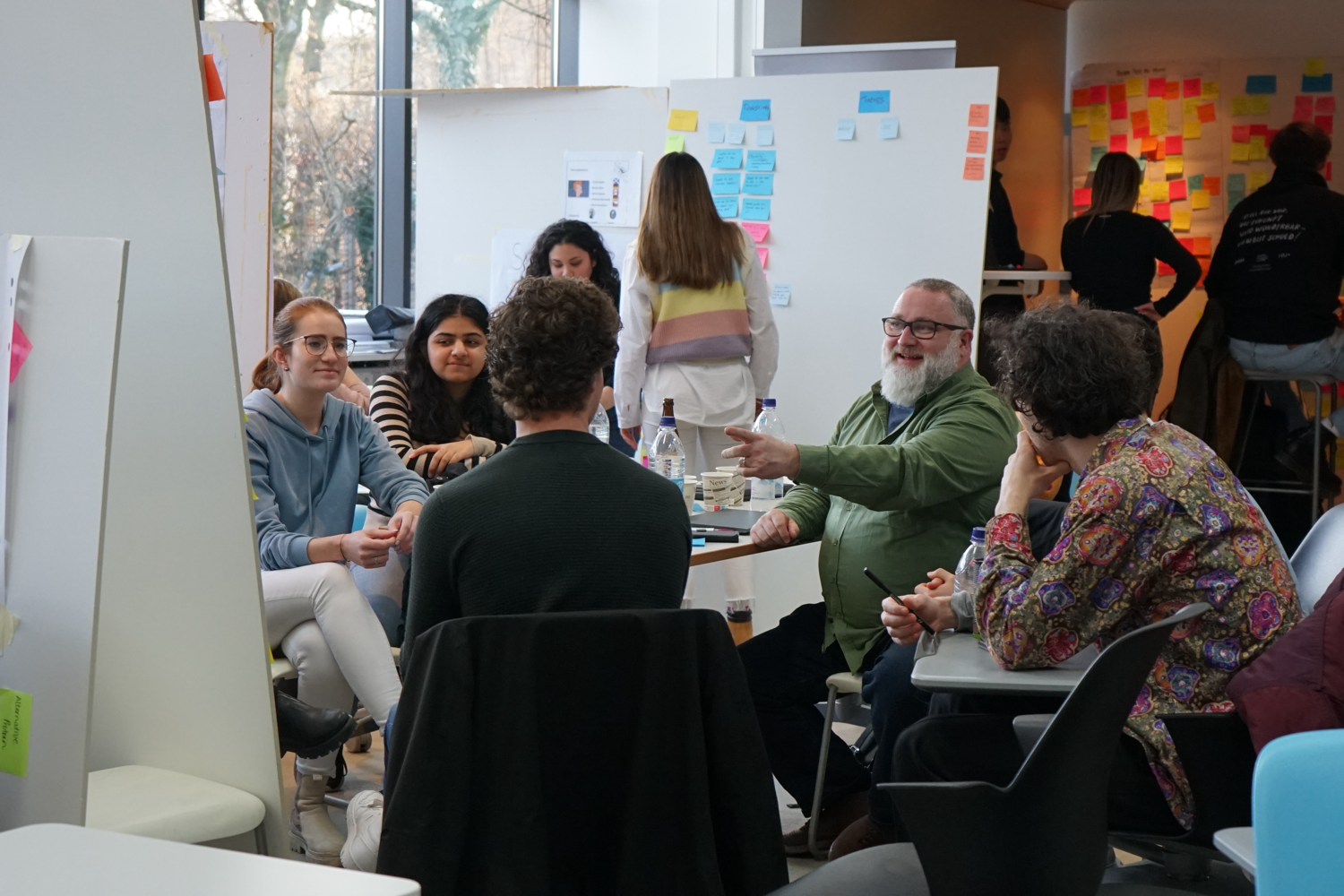 A diverse group of people gathered in a modern, collaborative workspace with whiteboards and sticky notes, engaging in discussion.