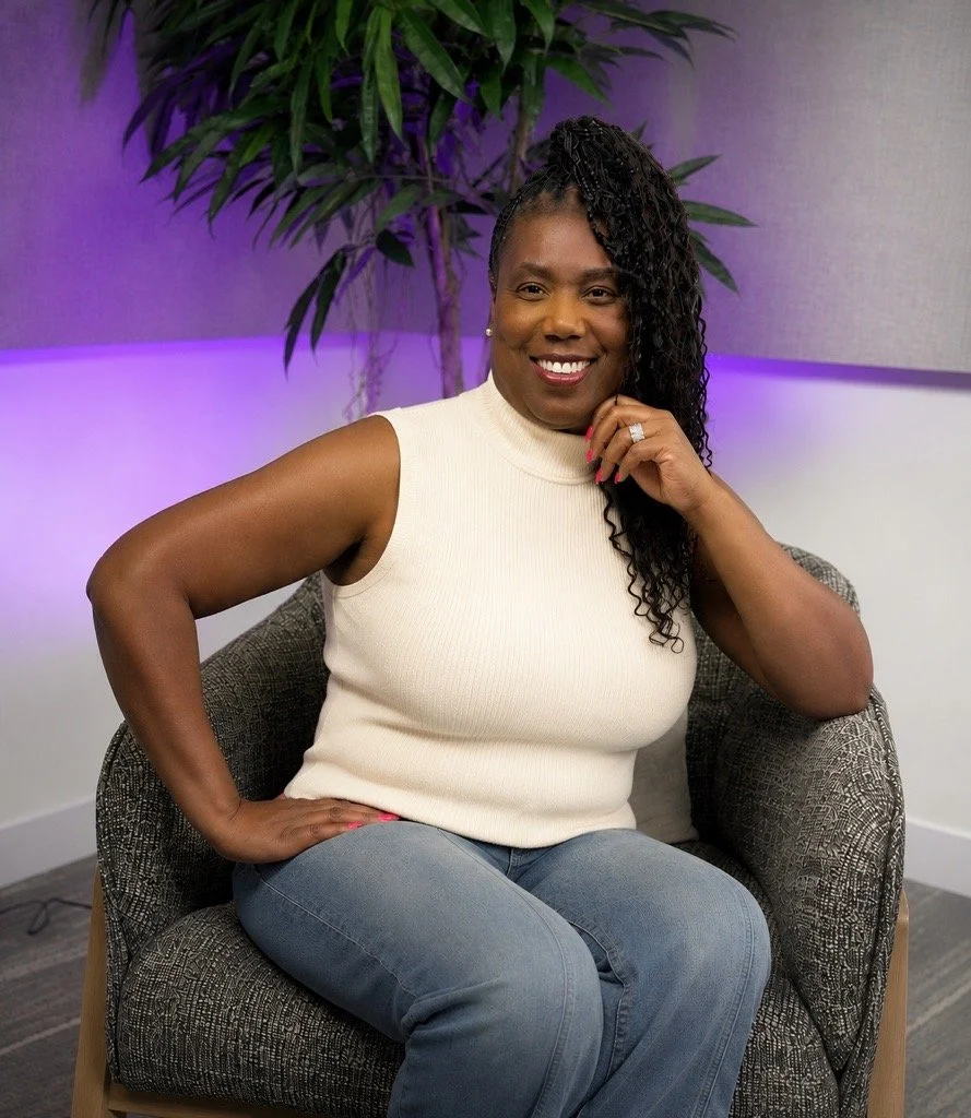 A smiling woman with dark curly hair, wearing a white sleeveless turtleneck sweater and blue jeans, sitting on a patterned armchair in front of a purple-lit plant background.