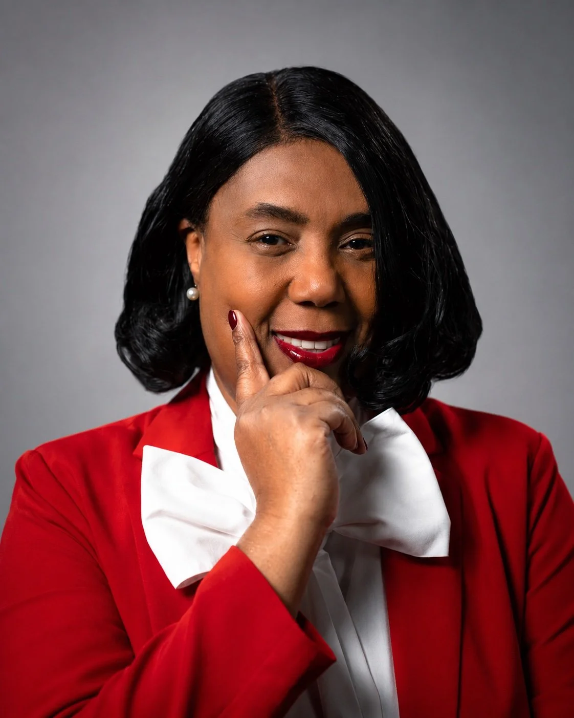 Portrait of an African American woman with black hair styled in loose curls, wearing a red blazer with a white shirt and pearl earrings, smiling confidently with her hand on her chin against a gray background.