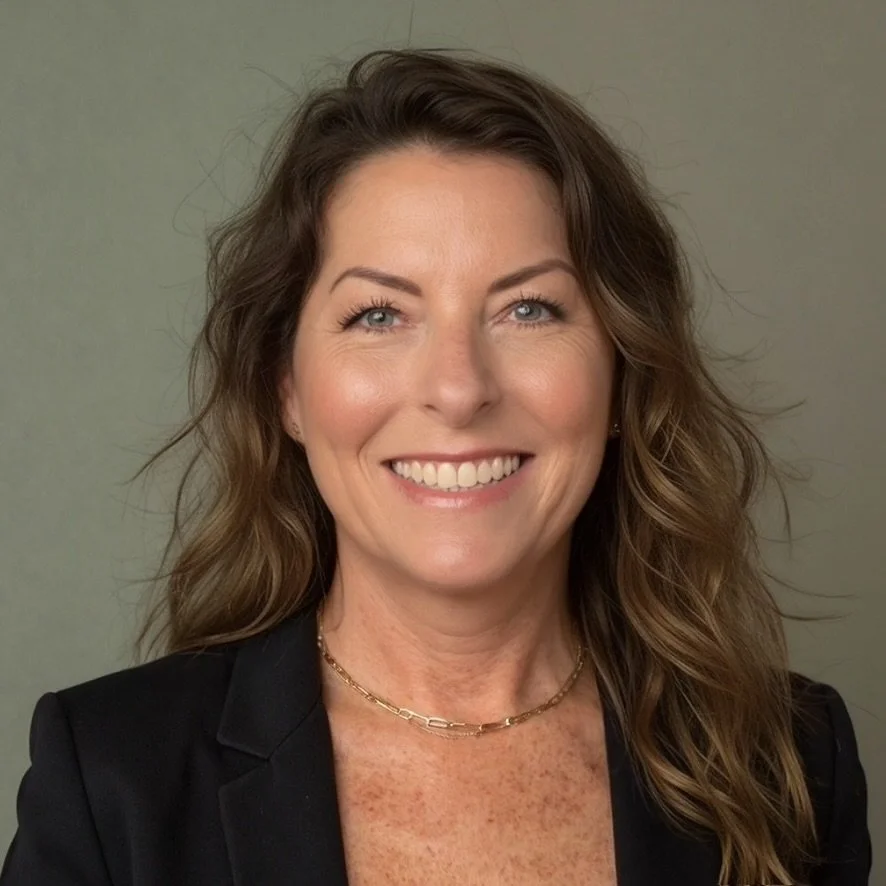 A smiling woman with wavy brown hair, wearing a black blazer and a gold chain necklace, against a plain gray background.