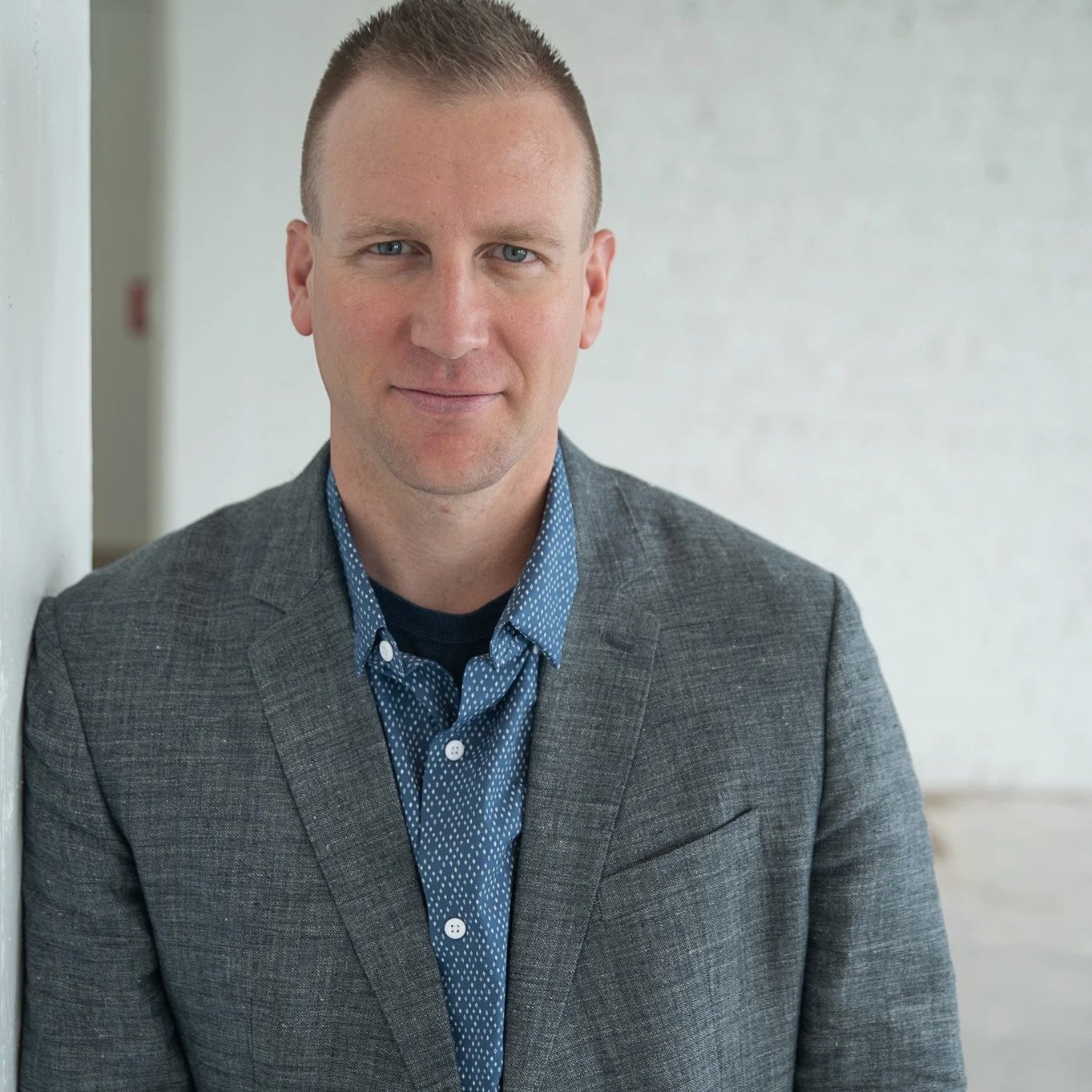 A man in a gray blazer and blue patterned shirt standing indoors against a light-colored wall, looking at the camera.
