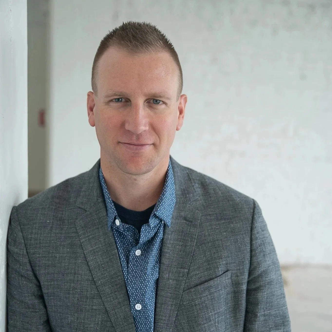 Portrait of a man with short hair, wearing a gray blazer and blue shirt with white dots, standing against a light-colored wall.