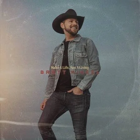 Man wearing a black cowboy hat and denim jacket, smiling and posing with one hand in pocket, against a blue background with partially visible text. Make a life Not a Livin. by Brett Kissel Juno Award Winning Country Album of the Year.
