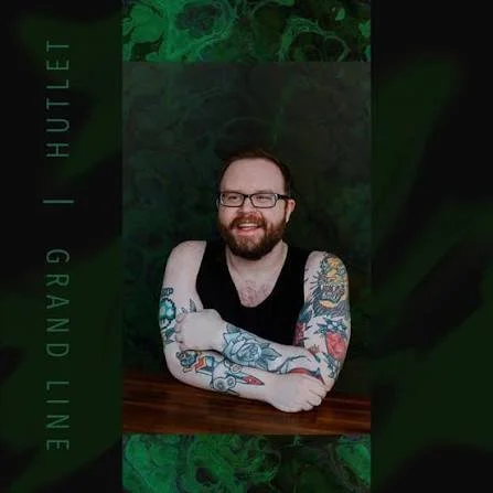 A man with glasses, a beard, and tattoos on his arms sitting at a wooden table, smiling, with a dark green or black marble background. Hutlet