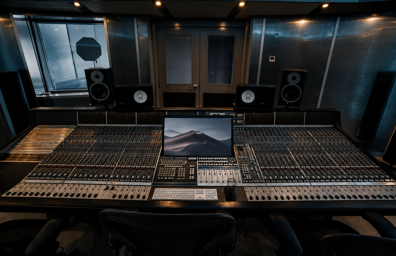 Professional recording studio with a large mixing console, computer monitor, studio speakers, and soundproof walls.