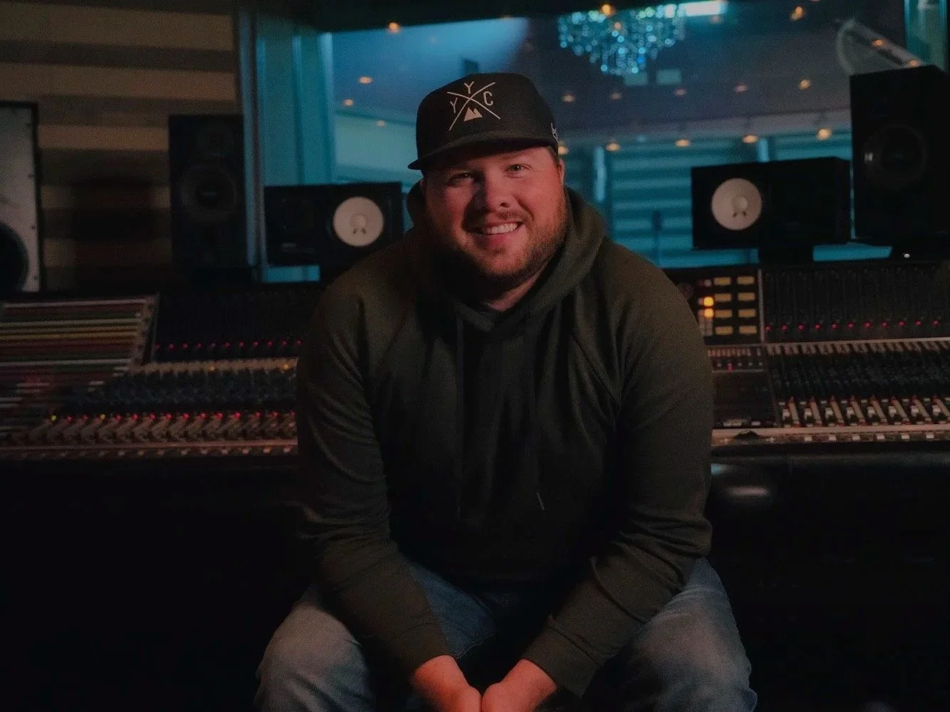A man wearing a hat and hoodie sitting in front of a music recording studio mixing console with speakers and a blue-lit room in the background. Brennan Kennedy JUNO and CCMA award winning Producer, Engineer.