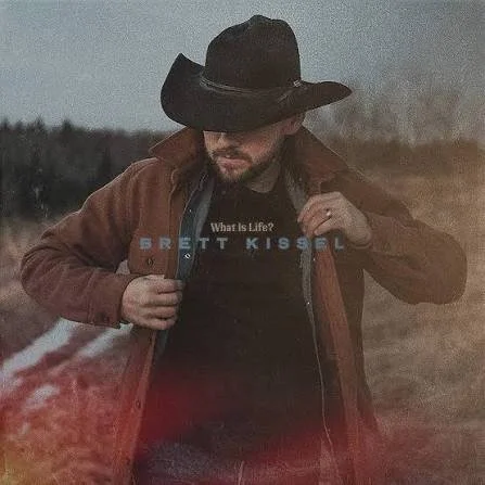 A man wearing a wide-brimmed black hat and a brown jacket, holding his jacket open to reveal a black t-shirt with the phrase "What is Life?" by Brett Kissel. JUNO Award Winning Country Album of the Year.