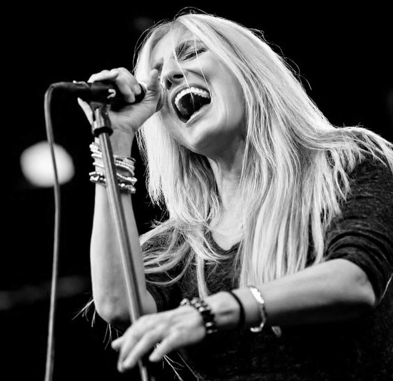 A woman singing passionately into a microphone during a performance, with long blonde hair and wearing bracelets. Sass Jordan