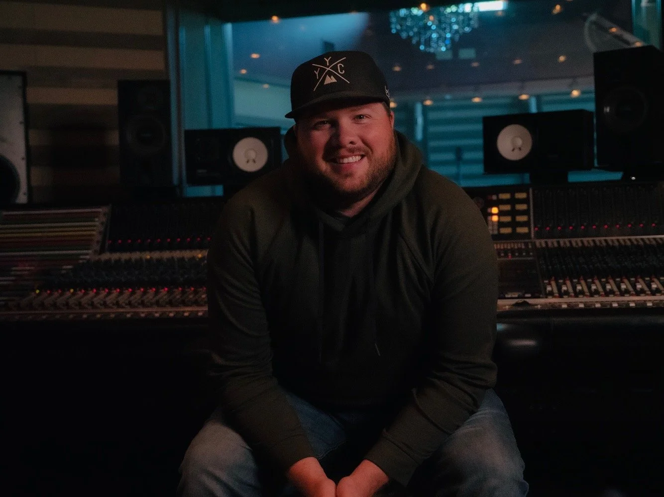 A smiling man sits in a music studio with sound equipment and speakers behind him, wearing a black hoodie and a cap with a logo.