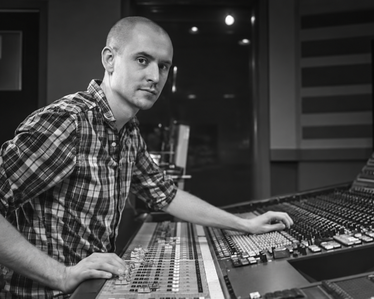 A man in a plaid shirt operating a large audio mixing console in a recording studio. Spencer Cheyne