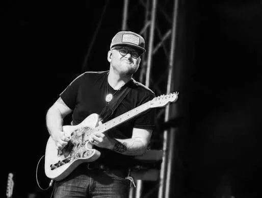 A man wearing a cap, glasses, and a black t-shirt playing an electric guitar on stage with stage lighting in the background. Matty McKay