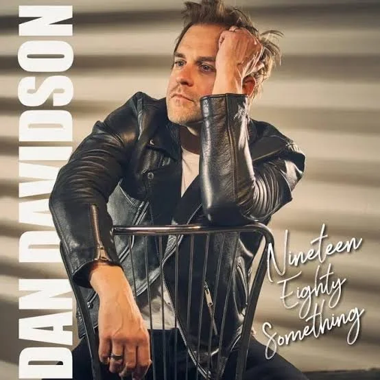 Man with styled hair and a beard wearing a black leather jacket, sitting with his hand on his head on a chair, with text reading "DAN DAVIDSON" and "Nineteen Eighty Something".