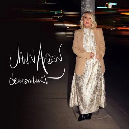 Woman with blonde hair wearing a beige blazer and cream-colored lace dress standing against a dark background with city lights in motion. Jann Arden