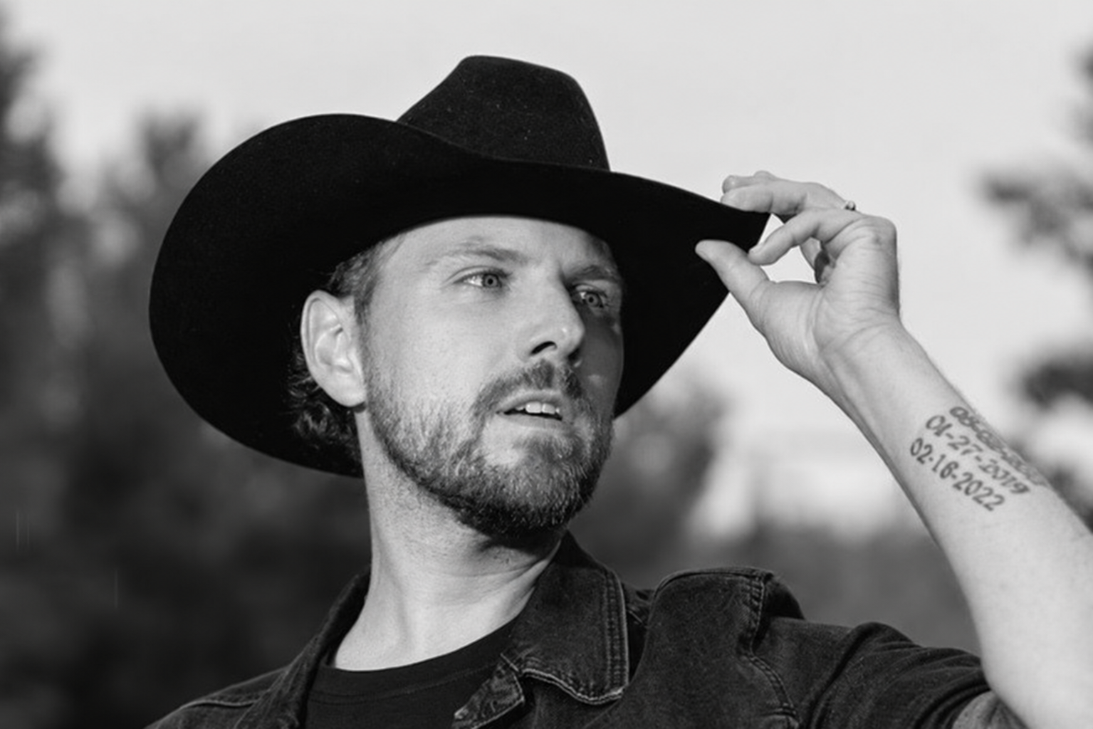 A man wearing a black cowboy hat and denim jacket looking into the distance, holding his hat with one hand, with a tattoo on his arm, in black and white. Brett Kissel