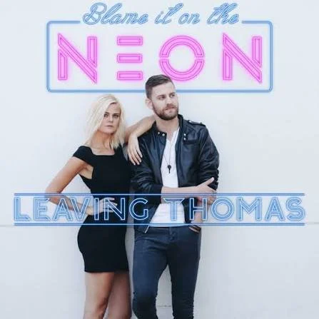 Two people, a woman and a man, standing in front of a neon sign that says "Blame it on the NEON, LEAVING THOMAS." The woman is wearing a black dress with short sleeves, and the man is wearing a black leather jacket and jeans.