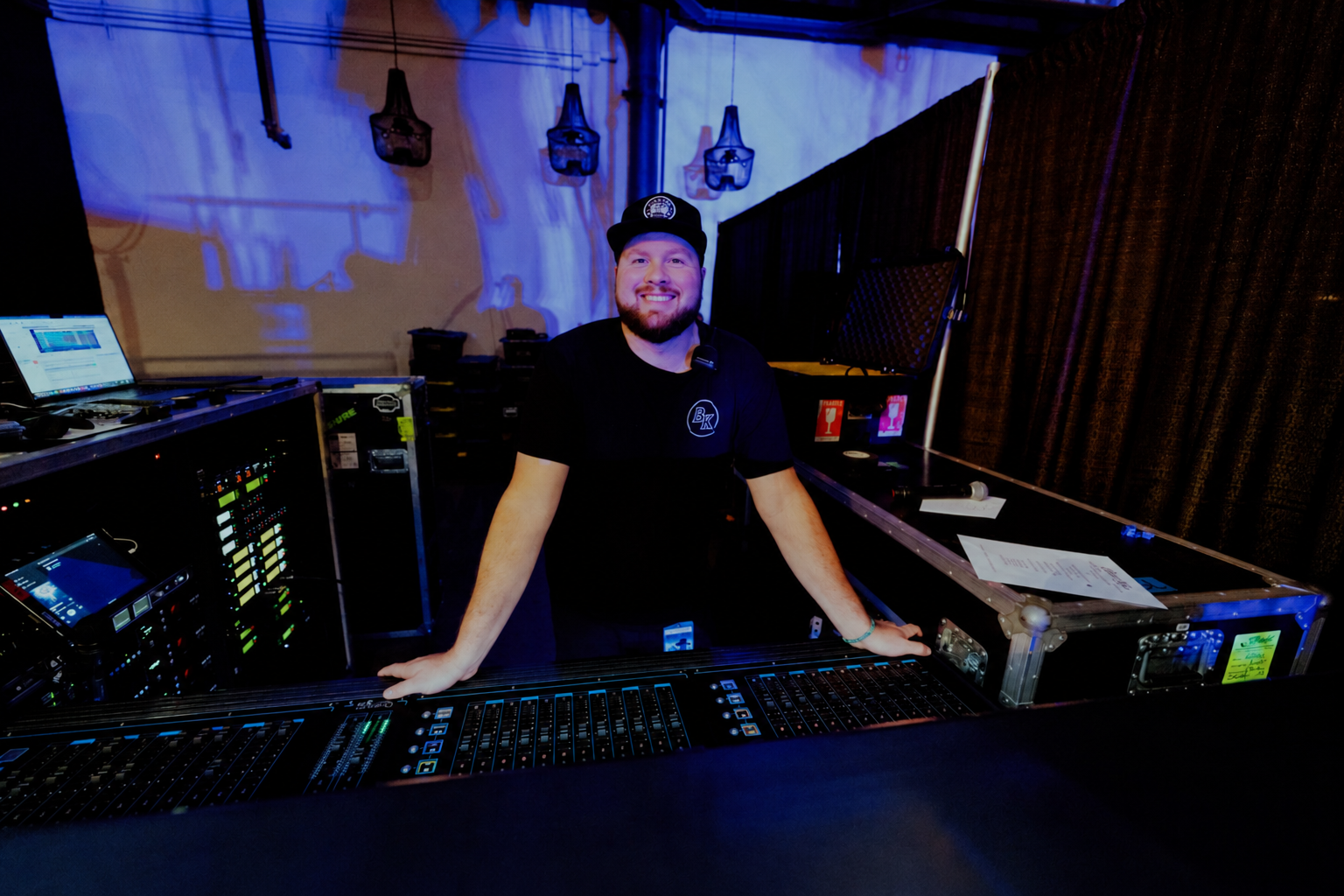 A smiling man with a beard, wearing a black shirt and a black cap, standing behind a DJ booth with equipment, in a dark room with blue lighting and hanging lights in the background.