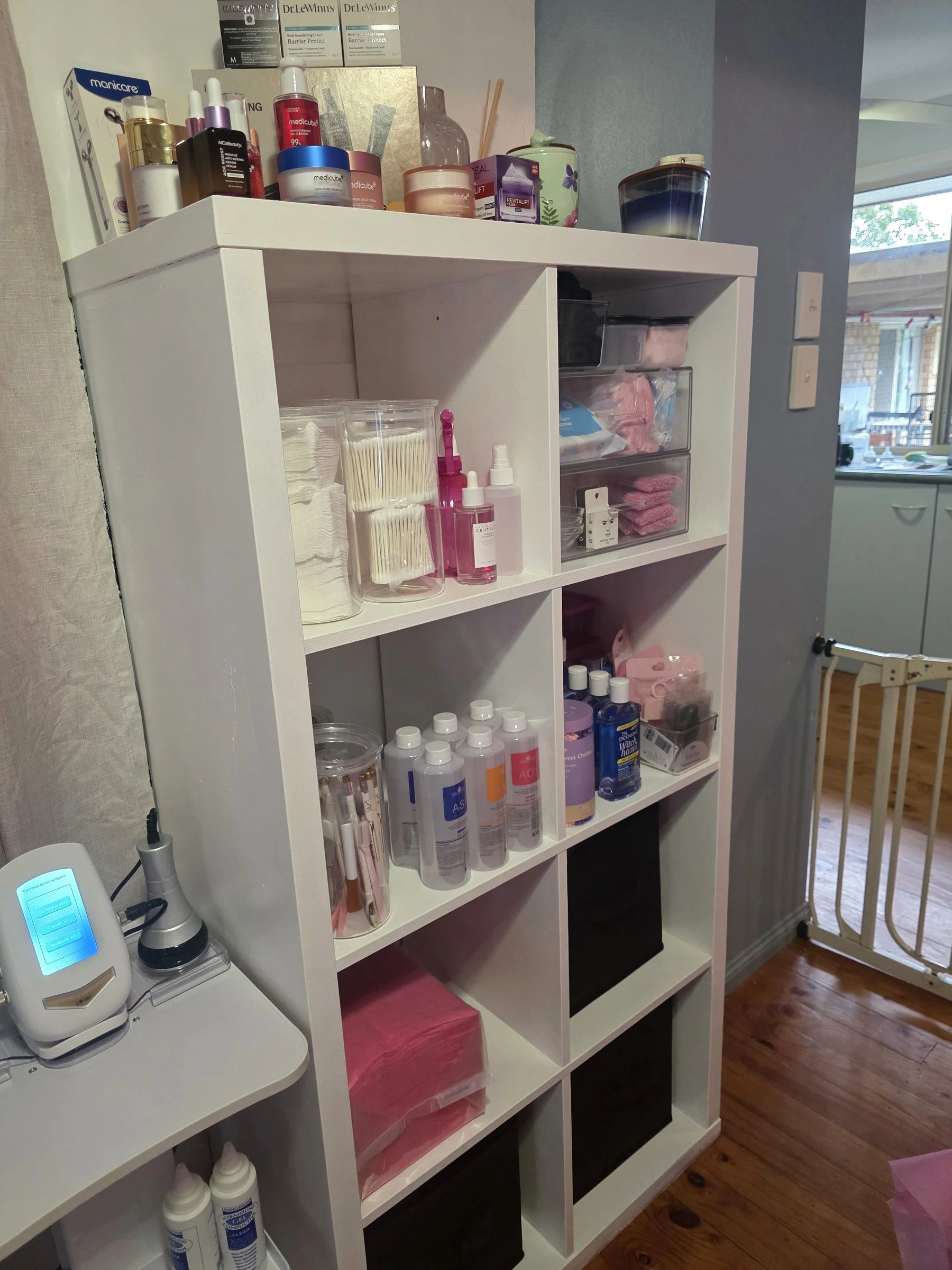 A white shelving unit filled with various skincare, beauty, and medical supplies, organized in clear and black containers with some bottles and jars on top and inside the shelves. A device is seen on the left side on a nearby surface.