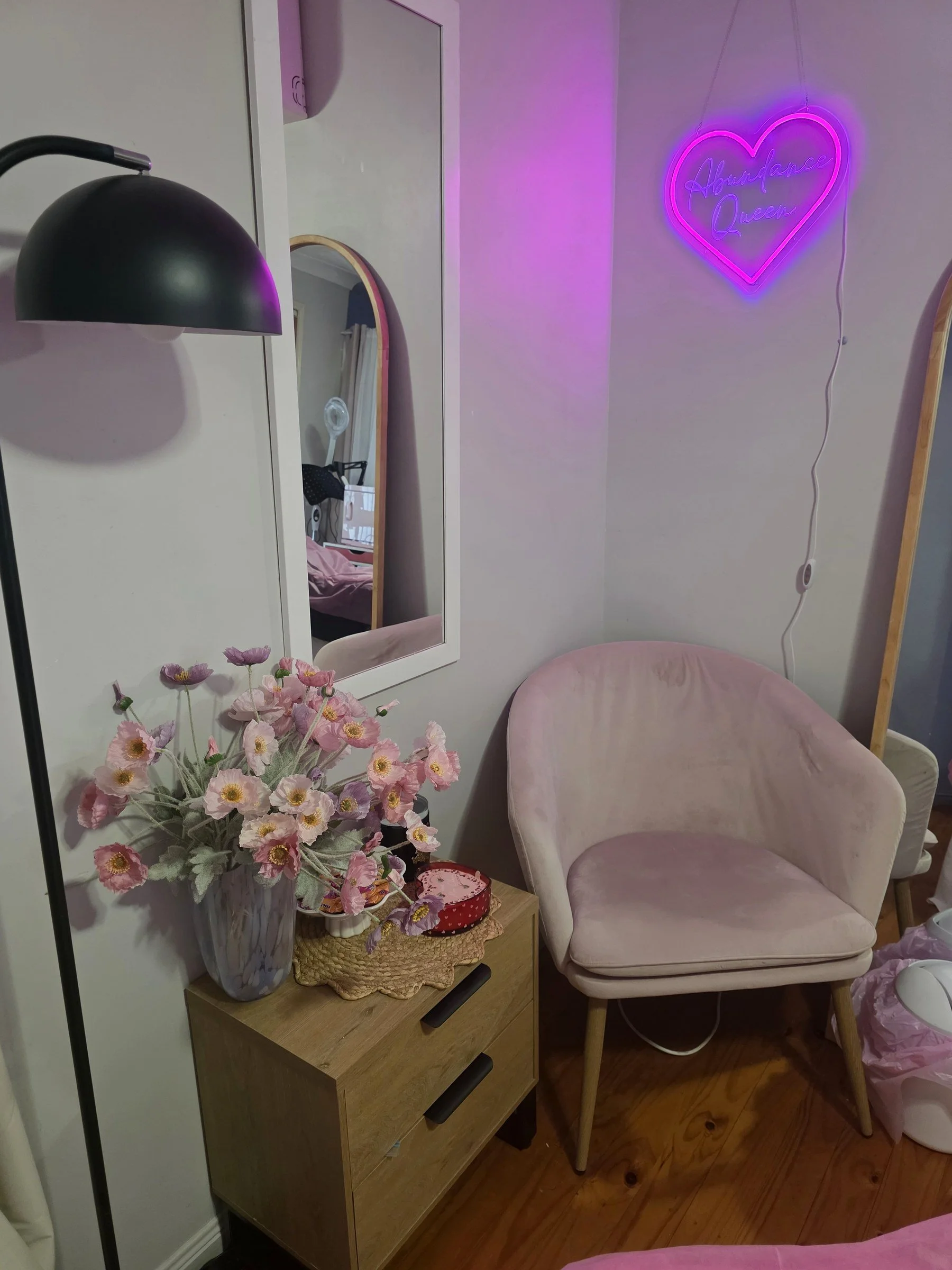 A cozy corner of a room featuring a pink velvet armchair, a wooden side table with a woven doily, a pink flower arrangement in a tall vase, a mirror on the wall reflecting part of the room, a black wall sconce, and a pink neon sign that reads 'Abundance Queen' in a heart shape. The floor is wooden, and there's a pink plastic bag on the floor next to the chair.