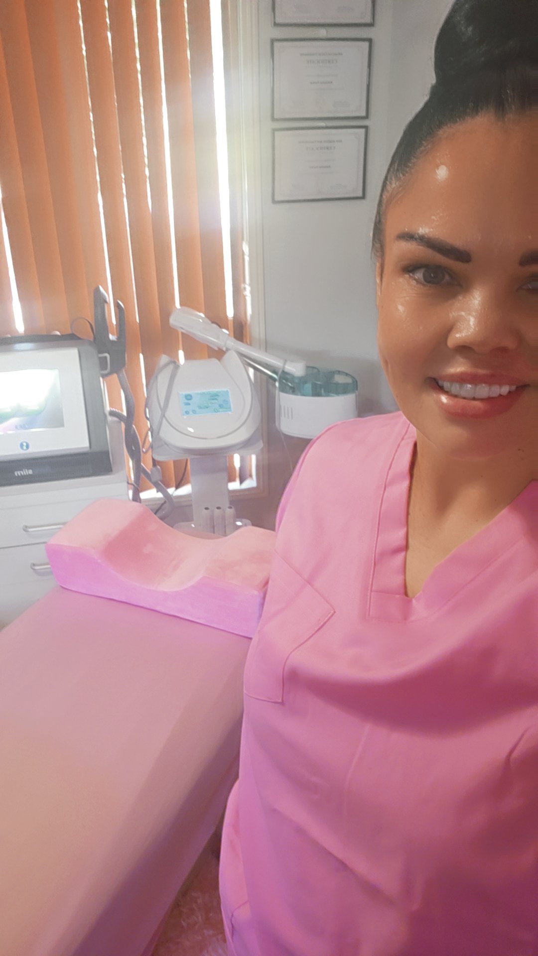 A woman in pink scrubs smiling in a medical or spa treatment room, with equipment and certificates on the wall behind her.