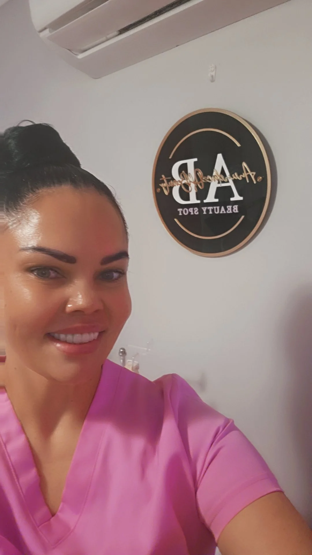 A woman in pink scrubs taking a selfie in front of a wall with a round sign that reads 'Aesthetic Beauty Spot'.