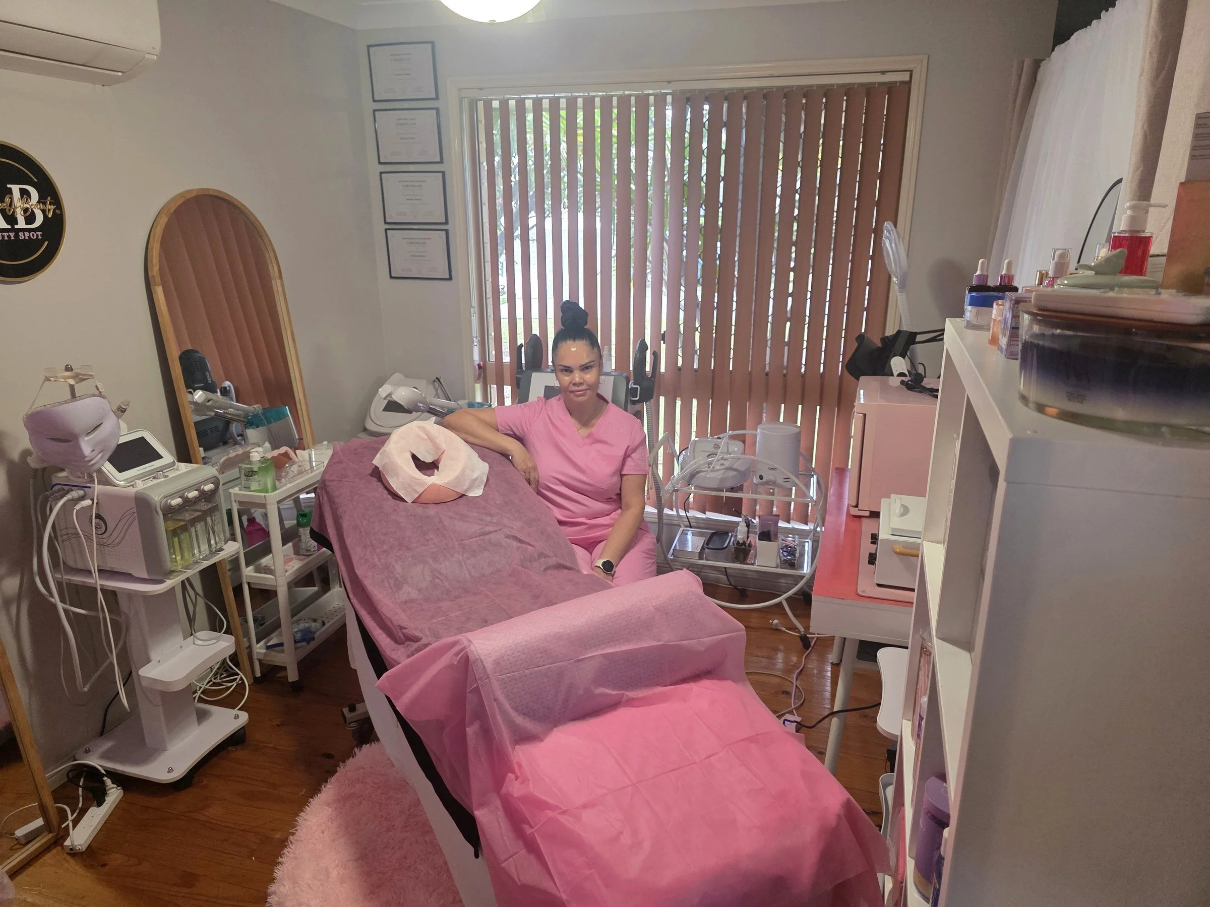 A woman in pink scrubs sitting on a treatment bed in a medical or spa room, surrounded by medical equipment and supplies, with certificates on the wall and a window with wooden blinds.