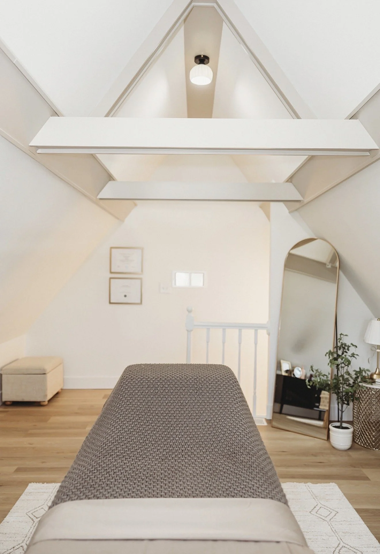 Cozy attic bedroom with vaulted white ceiling, exposed beams, upholstered bed, wood floors, arched mirror, and potted plant