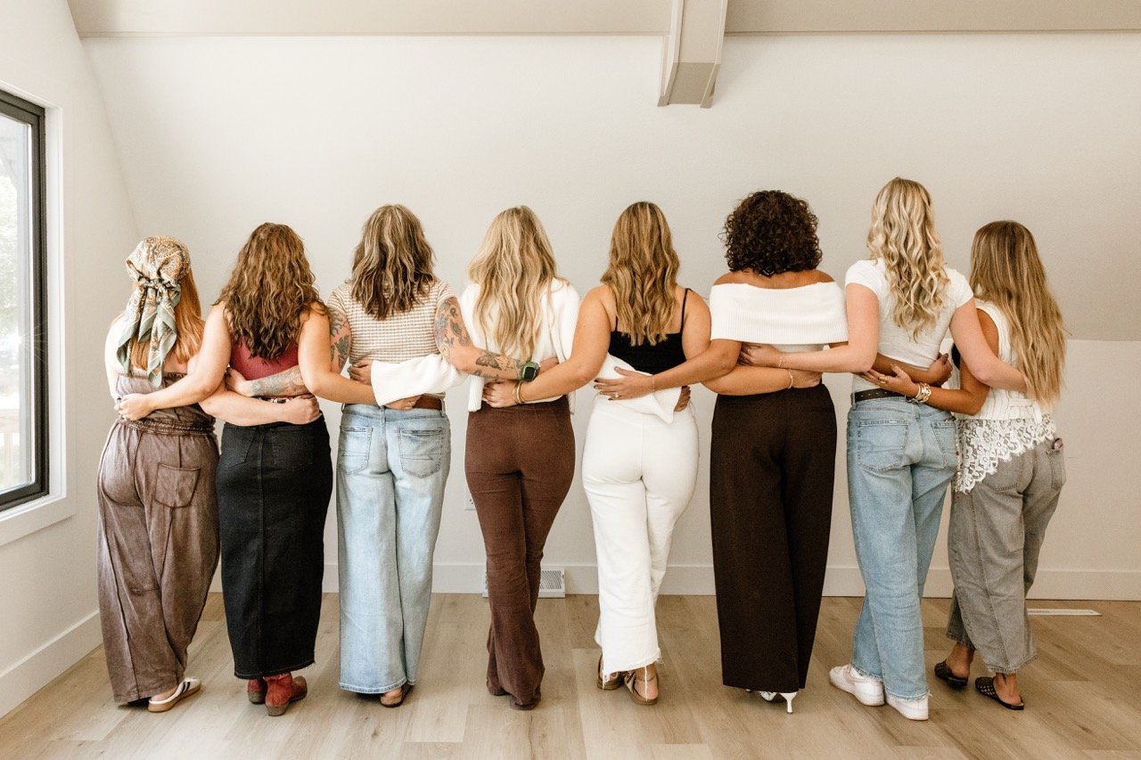 Group of eight women standing arm in arm with their backs to the camera in a bright room