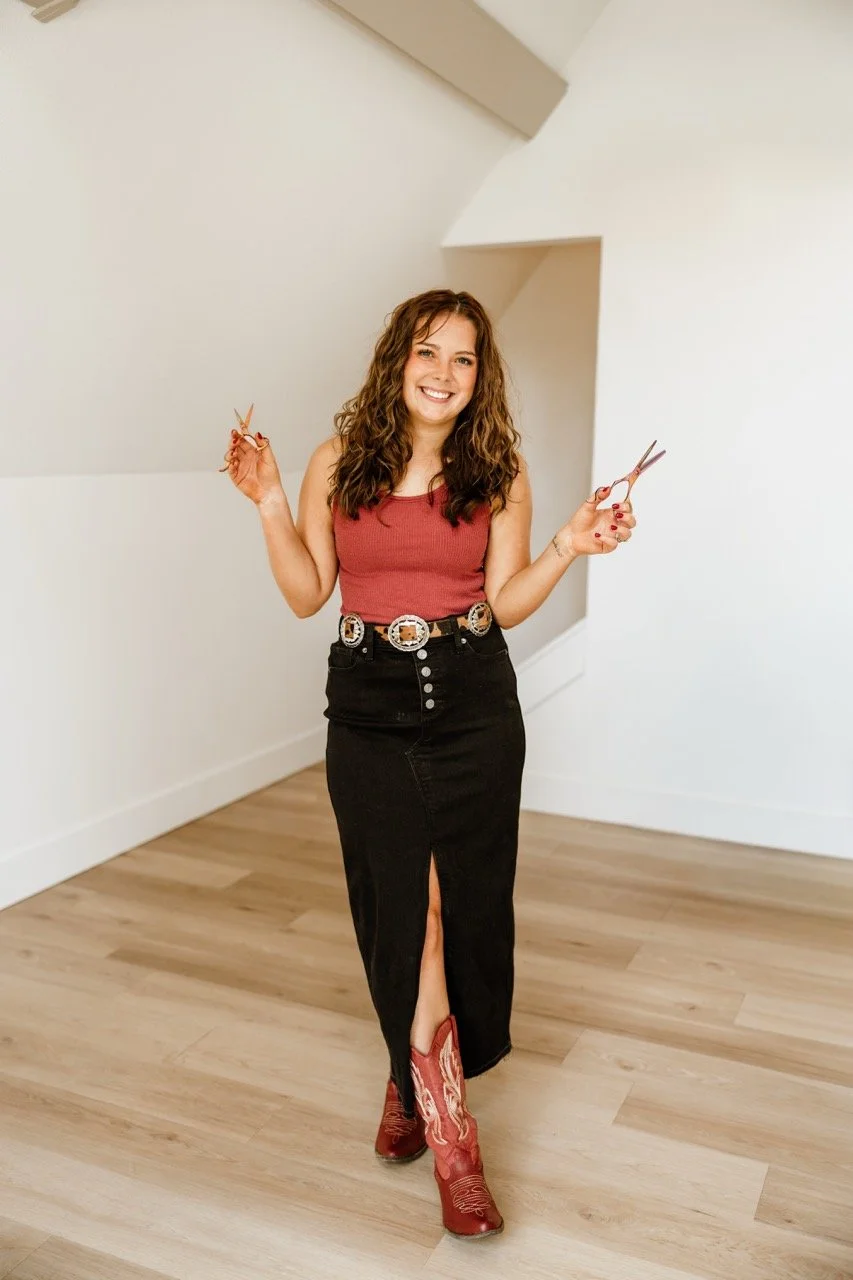 Smiling woman holding hair-cutting scissors while standing indoors in a red top, black skirt, and red cowboy boots