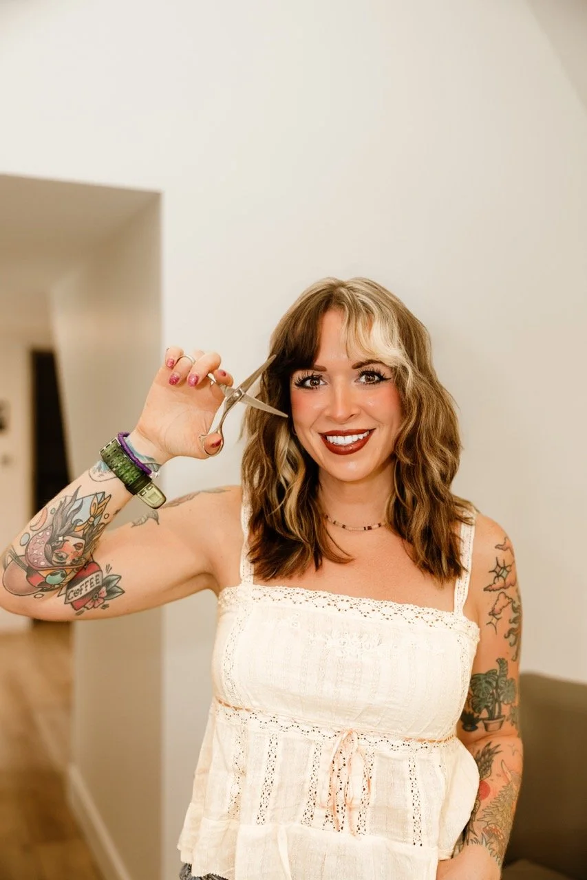 Smiling tattooed woman in a white top holding scissors near her hair