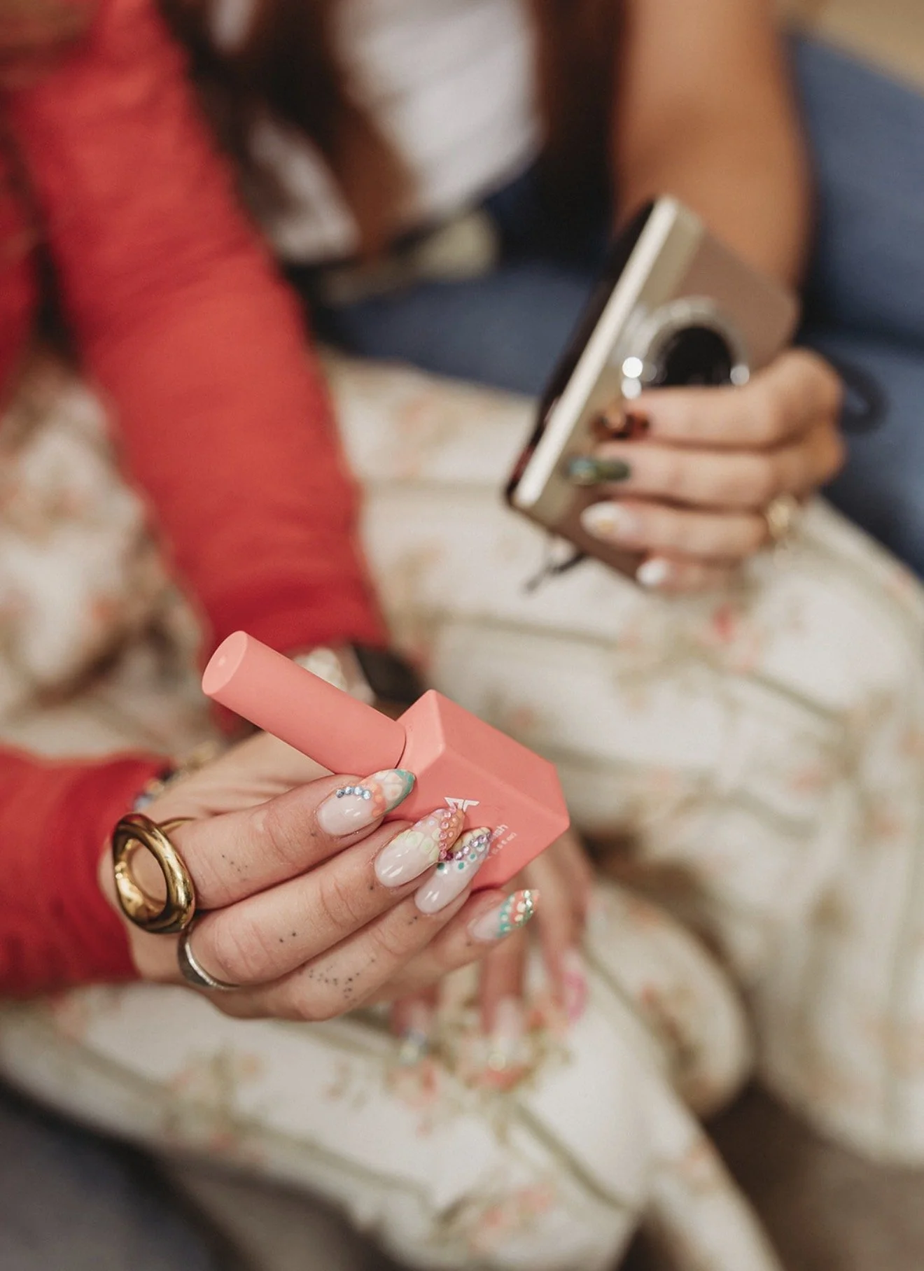 Close-up of a person with elaborate nail art holding a pink disposable vape while another person uses a smartphone in the background