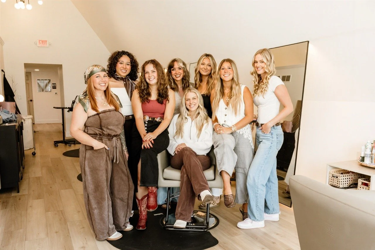 Group of eight women posing together in a bright salon or studio.