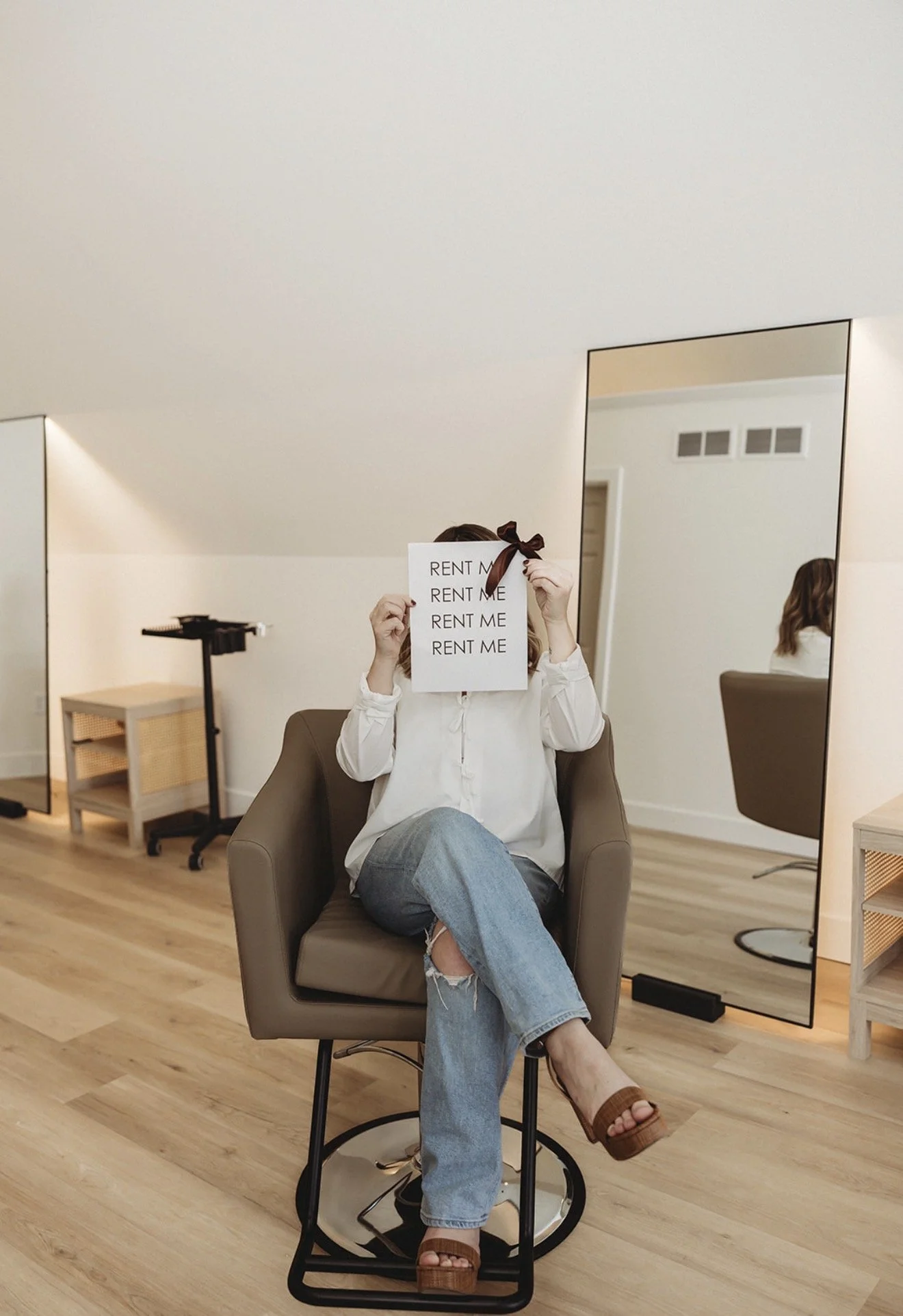 Woman sitting in a salon chair holding a sign reading “RENT ME” while wearing a burgundy bow.